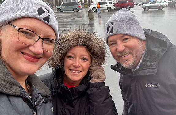 Group of three KELLYBRADY team members standing together outside on a rainy day, dressed in cold‑weather clothing.